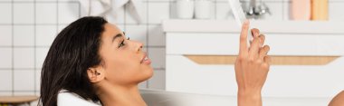 Genç Afrikalı Amerikalı kadın banyo yapıyor ve banyoda dijital tablete bakıyor.