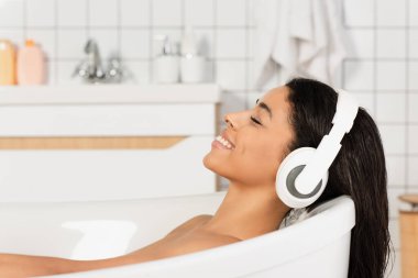Küvette gözleri kapalı, kulaklıkla müzik dinleyen mutlu genç bir kadın.