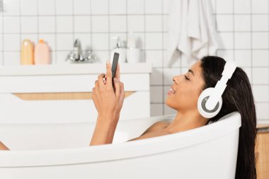 Kulaklıkla müzik dinleyen ve banyoda cep telefonuna bakan gülümseyen genç bir kadının yan görüntüsü