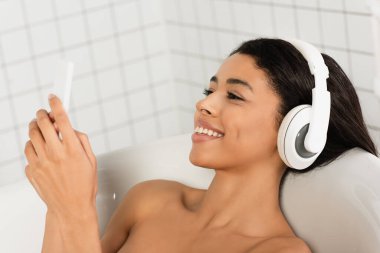 Kulaklıkla müzik dinleyen gülümseyen genç bir kadın banyoda cep telefonuna bakıyor.