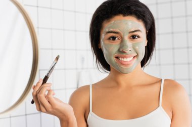 Yüzünde çamur maskesi olan gülümseyen Afrikalı Amerikalı kadın kozmetik fırça tutuyor ve banyoda kameraya bakıyor.