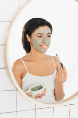 Gülümseyen genç Afrikalı Amerikalı kadın banyoda aynanın yanında makyaj fırçasıyla maske takıyor.