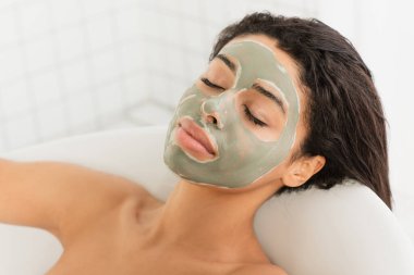 relaxed young african american woman with mud mask on face lying in bathtub clipart