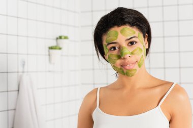 Yeşil maskeli genç Afro-Amerikan kadın banyoda kameraya bakıyor.