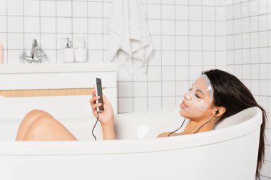 Yüzünde çarşaf maskesi olan, kulaklıkları ve cep telefonuyla küvette yatan genç bir kadın.
