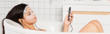 Yüzünde çarşaf maskesi olan, kulaklıkları ve cep telefonuyla küvette yatan Afro-Amerikan bir kadın.