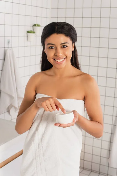 若いアフリカ系アメリカ人の女性が指で瓶の中のクリームにタオルを巻きバスルームのカメラを見て — ストック写真