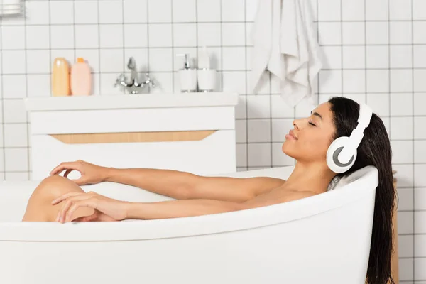 young woman with closed eyes resting and listening music in headphones in bathroom