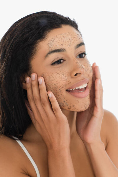 улыбающаяся молодая африканская американка, касающаяся лица скрабом, изолированным на белом