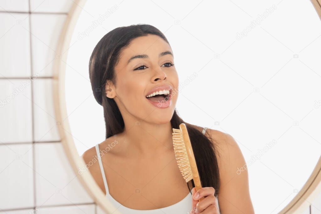 young african american woman singing with hair brush near mirror in bathroom