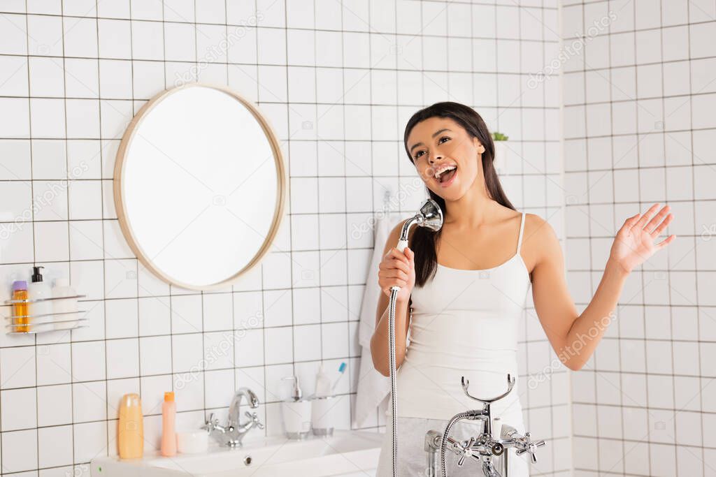 playful young african american woman in white clothes singing with showerhead in hand in bathroom