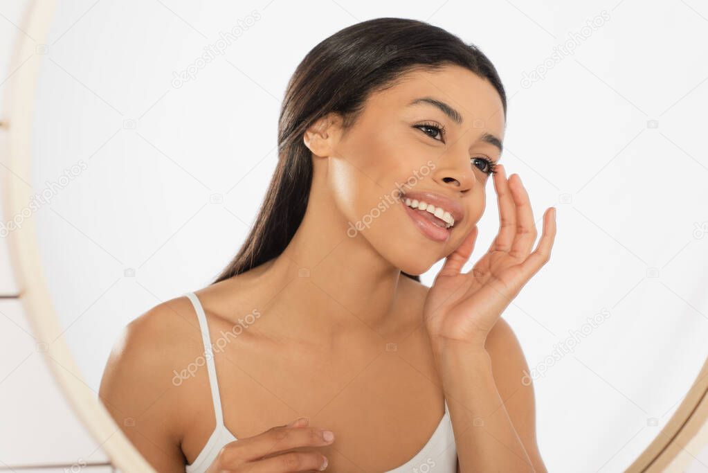 smiling young african american woman looking in mirror and touching face with hand in bathroom