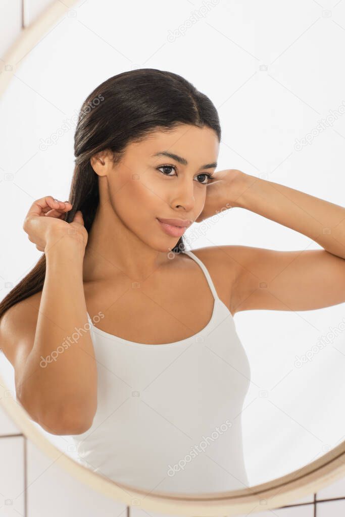 confident young african american woman looking in mirror and making hairstyle with hands in bathroom