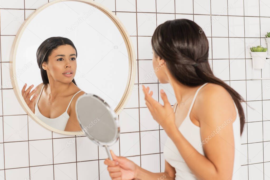 stressed young african american woman looking in mirror and holding hand near face in bathroom