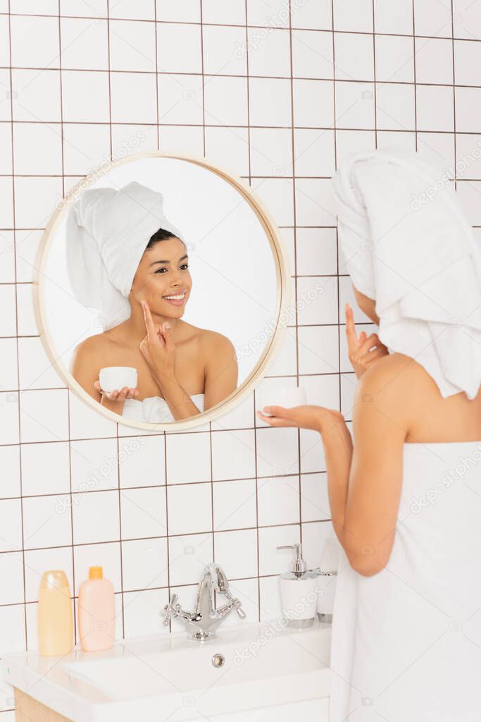 young african american woman wrapped in towels applying cream on face near mirror in bathroom