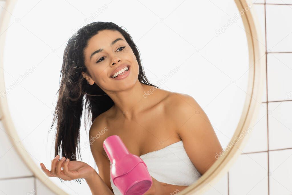 smiling young african american woman wrapped in towel styling hair with dryer near mirror in bathroom