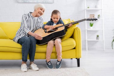 Kanepede dedenin yanında akustik gitar çalan gülümseyen çocuk. 