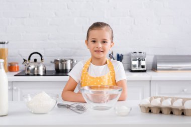 Önlüklü gülümseyen kız mutfak masasındaki yumurta, un ve sütün yanındaki kameraya bakıyor. 