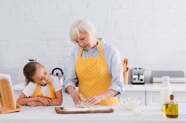 Kid looking at grandmother making dough near digital tablet and ingredients  clipart