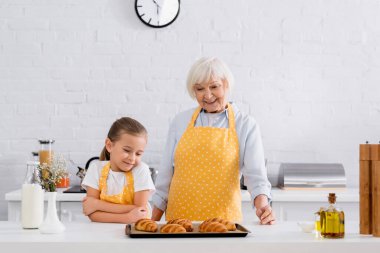 Cheerful granny and child looking at tasty croissants on kitchen table  clipart
