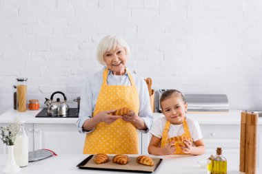 Smiling granny and kid holding tasty croissants near baking sheet in kitchen  clipart