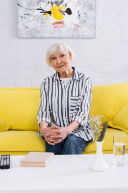 Gülümseyen yaşlı kadın kitapların, suyun ve uzaktan kumandanın yanındaki kameraya bakıyor. 