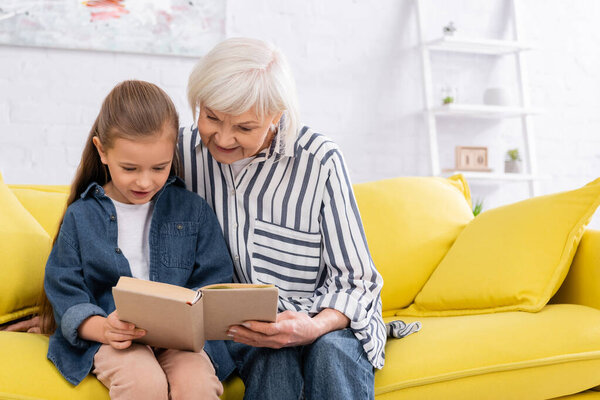 Улыбающаяся бабушка читает книгу с внучкой дома 