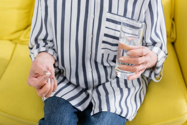 ソファの上で水の錠剤とガラスを保持しているシニア女性の作物ビュー — ストック写真