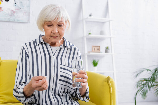 Upset elderly woman holding pill and glass of water in living room 