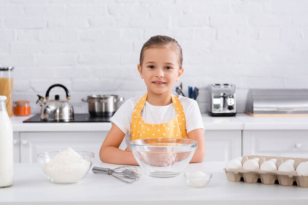 Smiling girl in apron looking at camera near eggs, flour and milk on kitchen table 