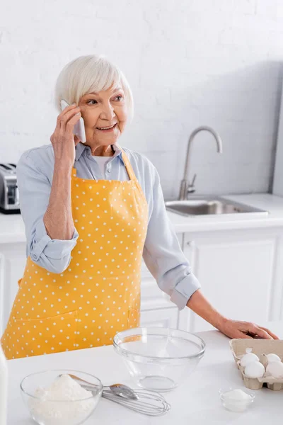 Ηλικιωμένη Γυναίκα Ποδιά Μιλάει Smartphone Κοντά Αλεύρι Και Αυγά — Φωτογραφία Αρχείου