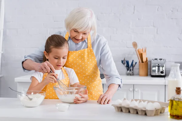 Granny and kid cooking together near ingredients in kitchen
