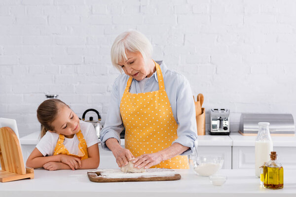 Kid looking at grandmother making dough near digital tablet and ingredients 