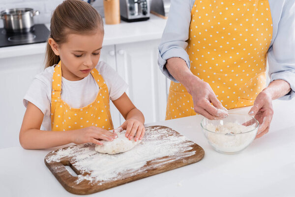 Kid making dough near granny with flour in kitchen 