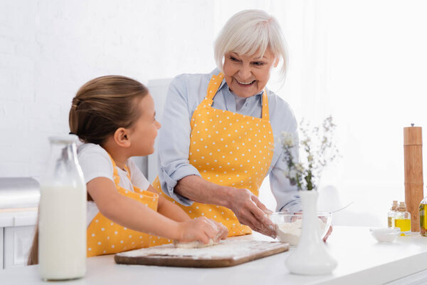 Cheerful granny holding flour and looking at kid with dough on blurred foreground in kitchen 