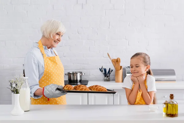 笑孩子在围裙看着奶奶与羊角面包在烘烤单 — 图库照片