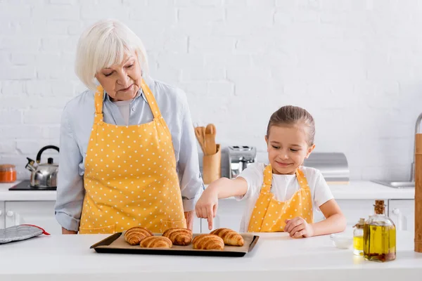 Chica Sonriente Delantal Apuntando Croissant Cerca Abuela Cocina — Foto de Stock