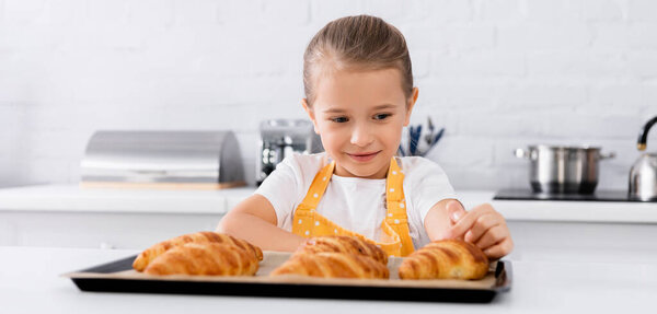 Smiling girl in apron touching baked croissant in kitchen, banner 