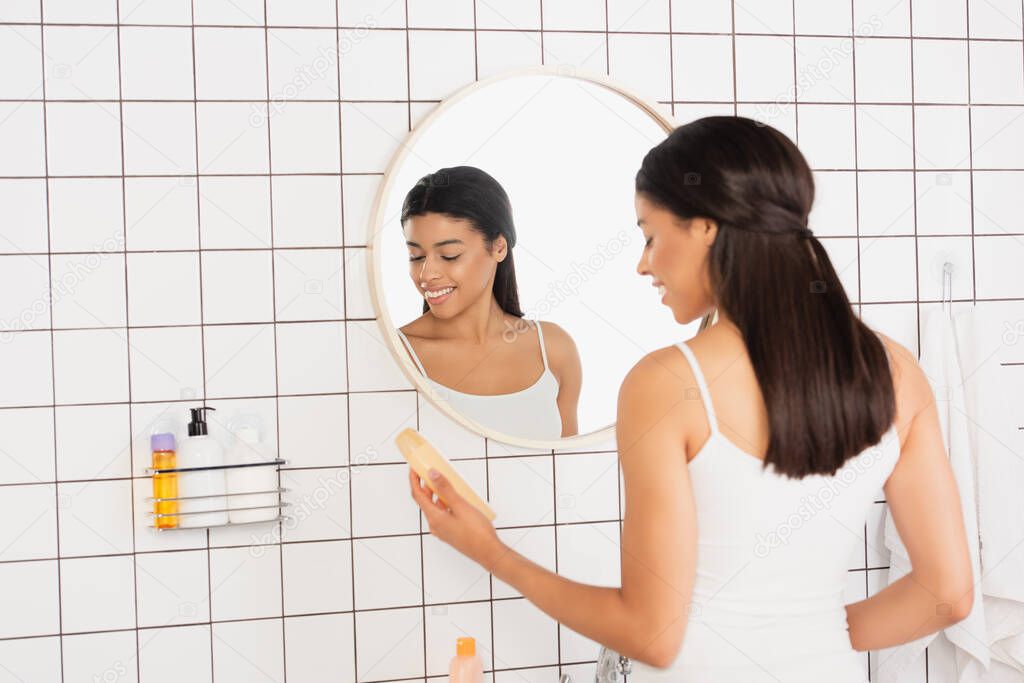young african american woman holding shower gel near mirror in bathroom