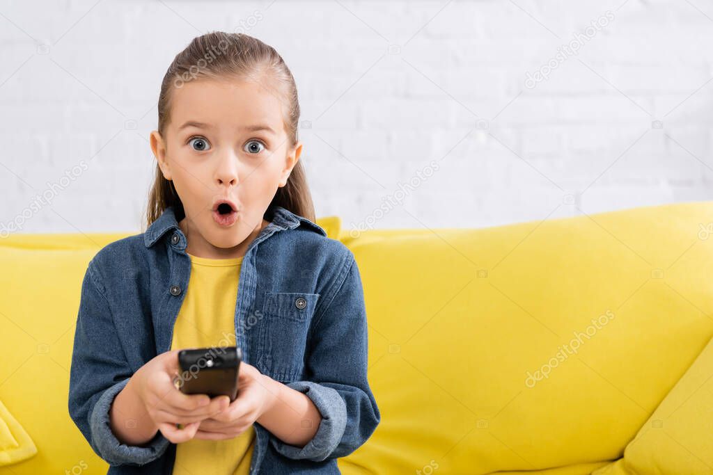 Shocked child holding blurred remote controller on couch 