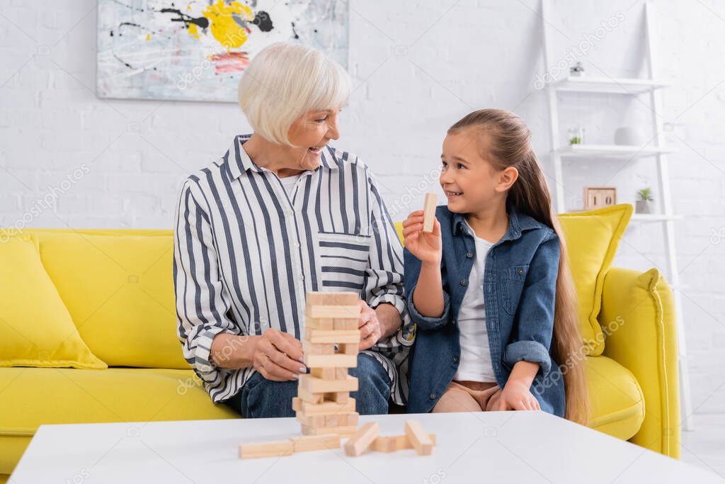 Smiling girl holding block of wood game near granny and tower on table 