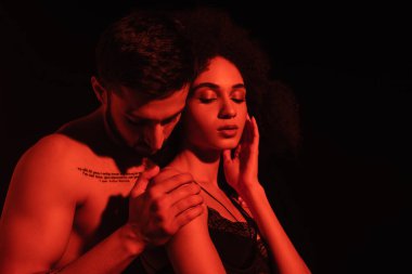 Siyah ışıkta izole edilmiş, Afro-Amerikan kız arkadaşının omzuna dokunan gömleksiz adam. 