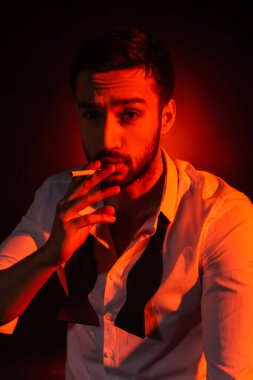 Bearded man in formal wear holding cigarette on black background with red lighting clipart