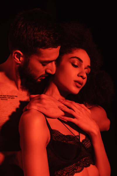 Shirtless man hugging sensual african american girlfriend in red light isolated on black 