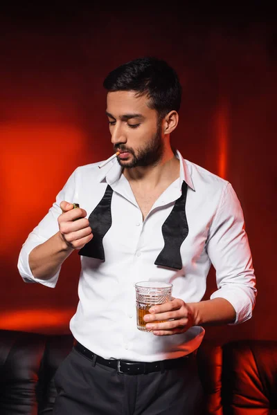 Bearded man with cigarette and whiskey holding lighter on black background with red lighting