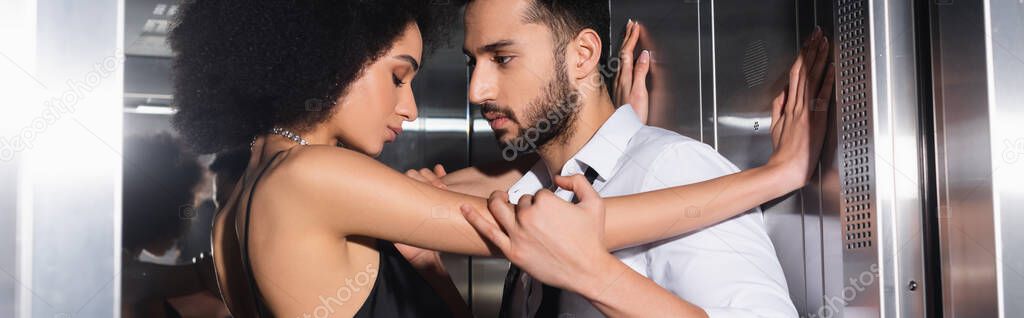 Side view of man in shirt touching african american girlfriend in elevator, banner 