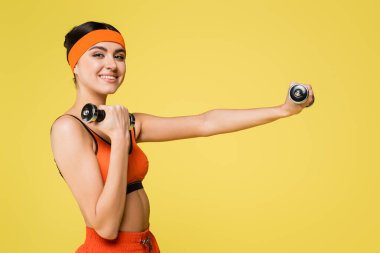 Sarı renkli dambıllarla çalışırken kameraya gülümseyen mutlu kadın.