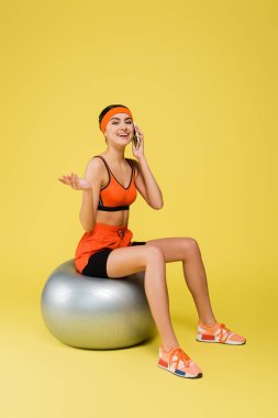 Sarı arka planda fitness topunun üzerinde otururken akıllı telefondan konuşan neşeli sporcu kadın.