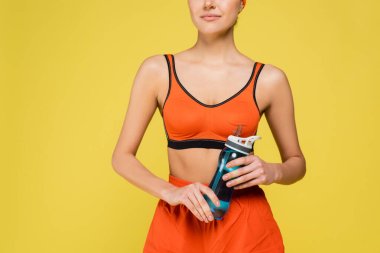 cropped view of woman in orange sportswear holding sports bottle isolated on yellow clipart