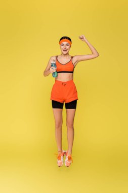 Spor şişesi atlayan neşeli sporcu kadın sarı renkte izole edilmiş zafer hareketi sergiliyor.